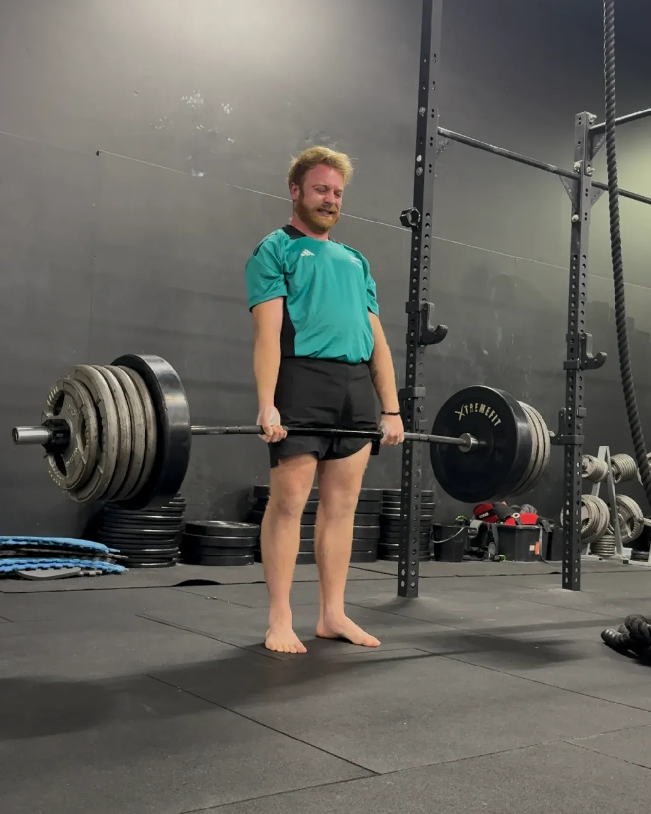 A member standing with a loaded barbell at his hips, barefoot on the gym floor