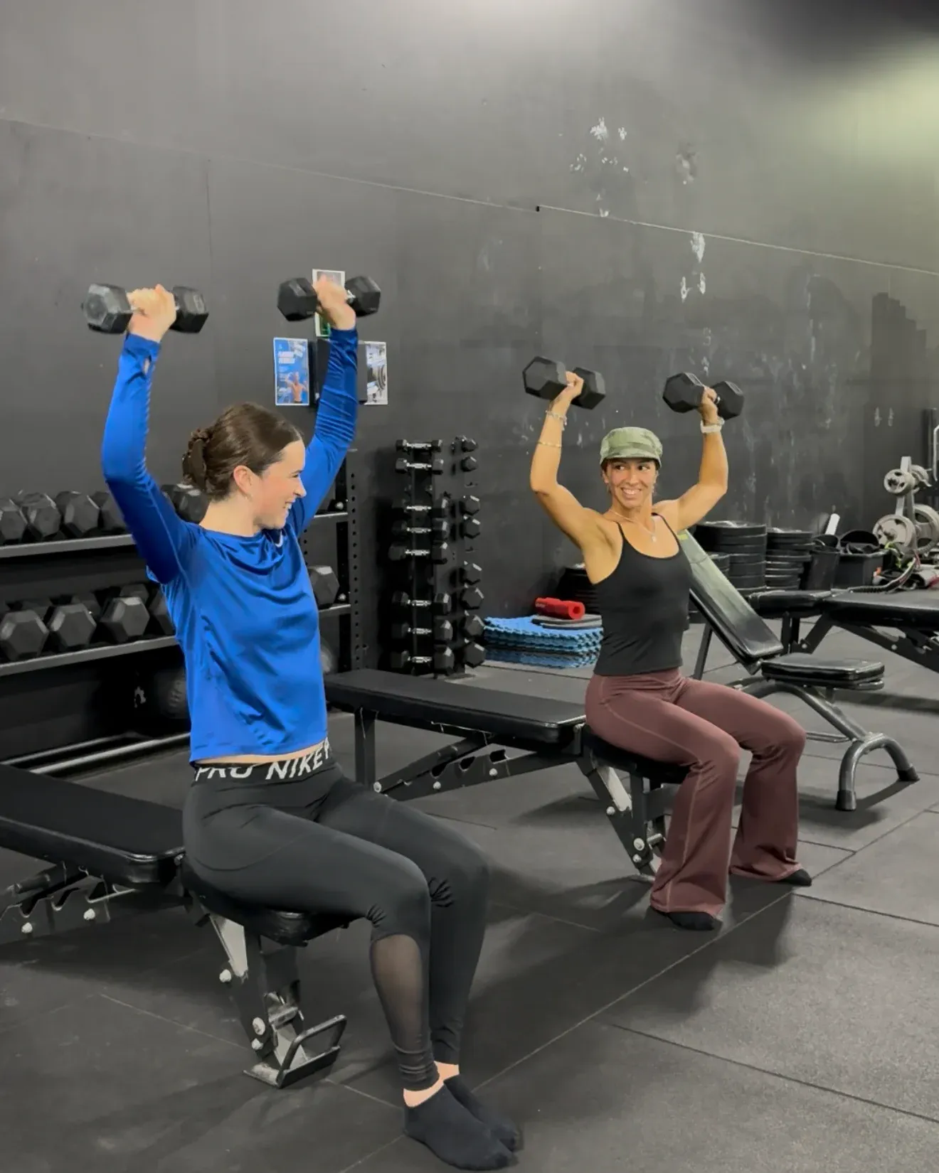 Two women seated on benches, pressing dumbbells overhead together