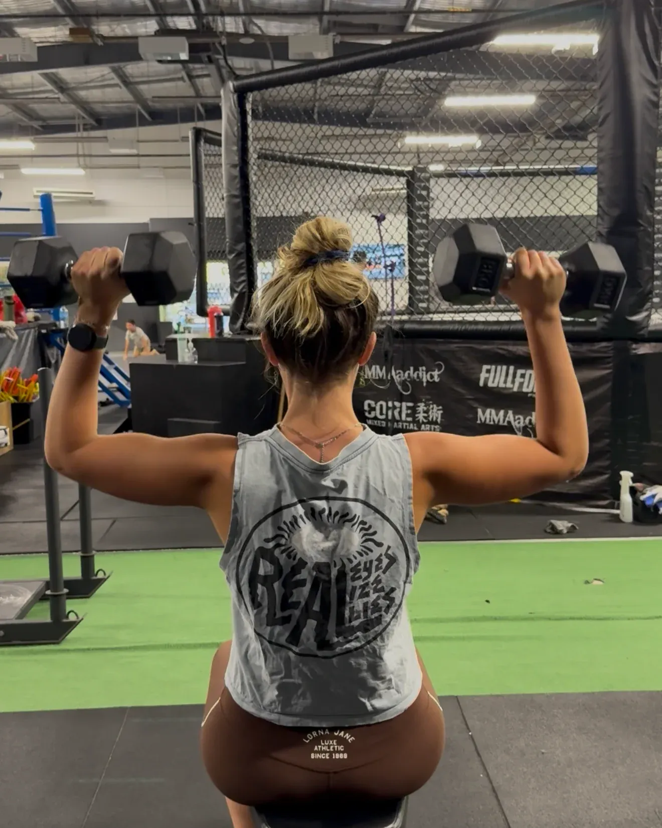 A member pressing dumbbells overhead on a bench, the CORE MMA cage behind her