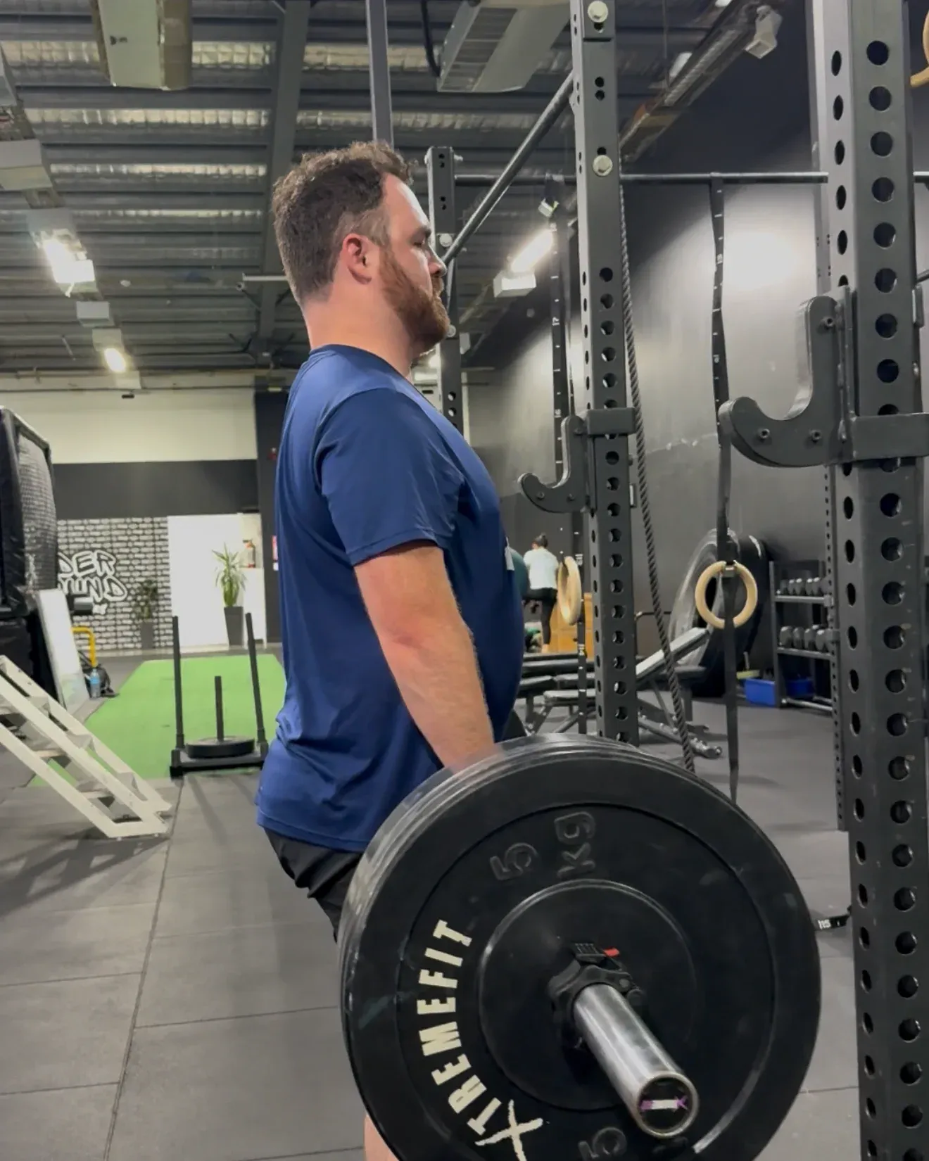A member holding a loaded barbell at the top of a deadlift beside the rack