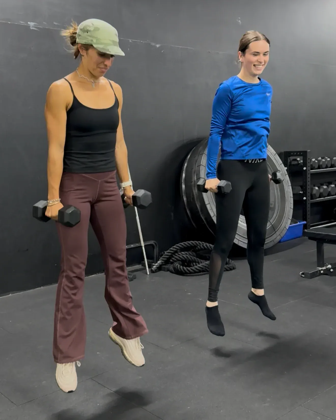 Two women mid-jump, dumbbells in hand, during a conditioning set
