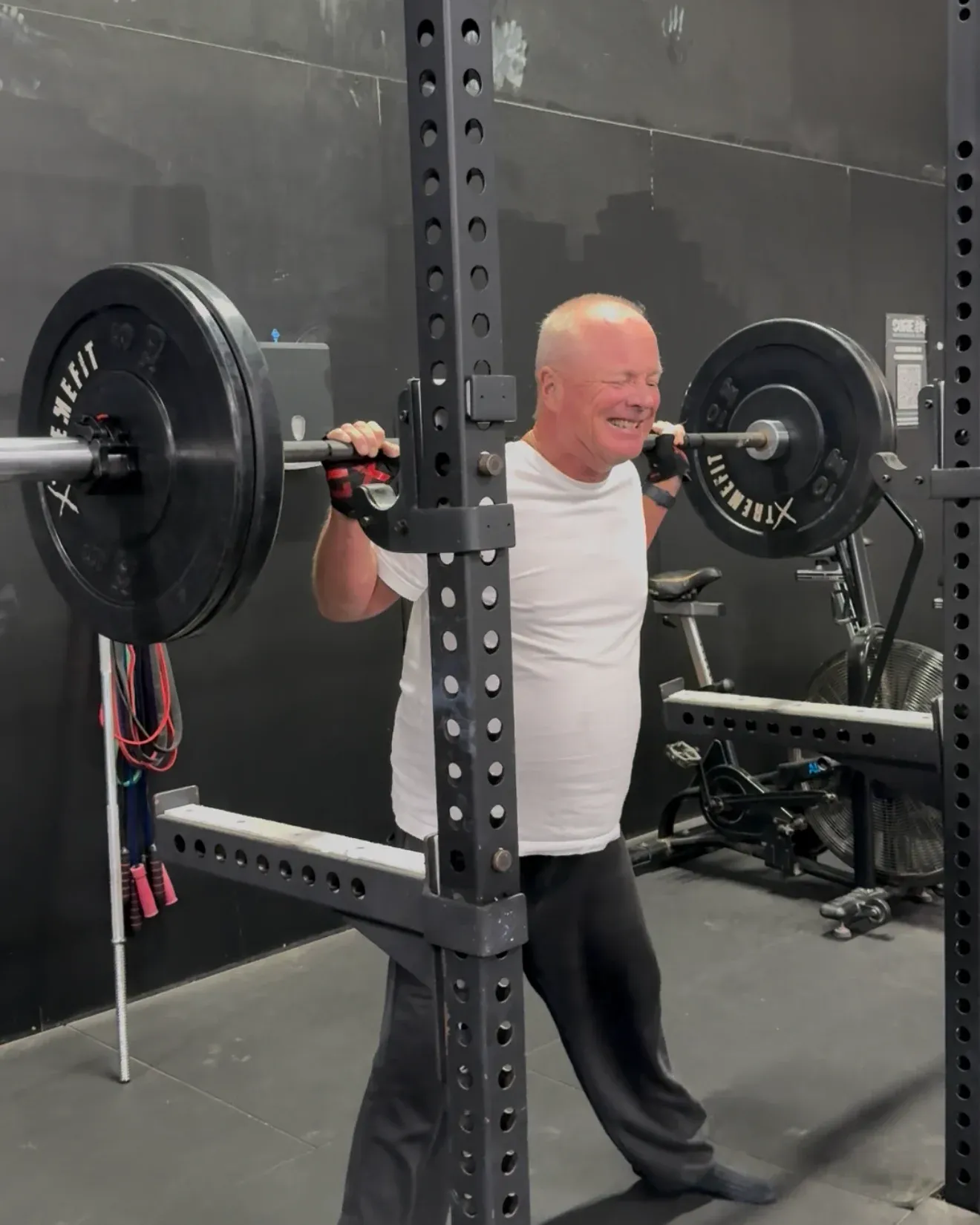 An older member grinning with a barbell across his shoulders in the squat rack