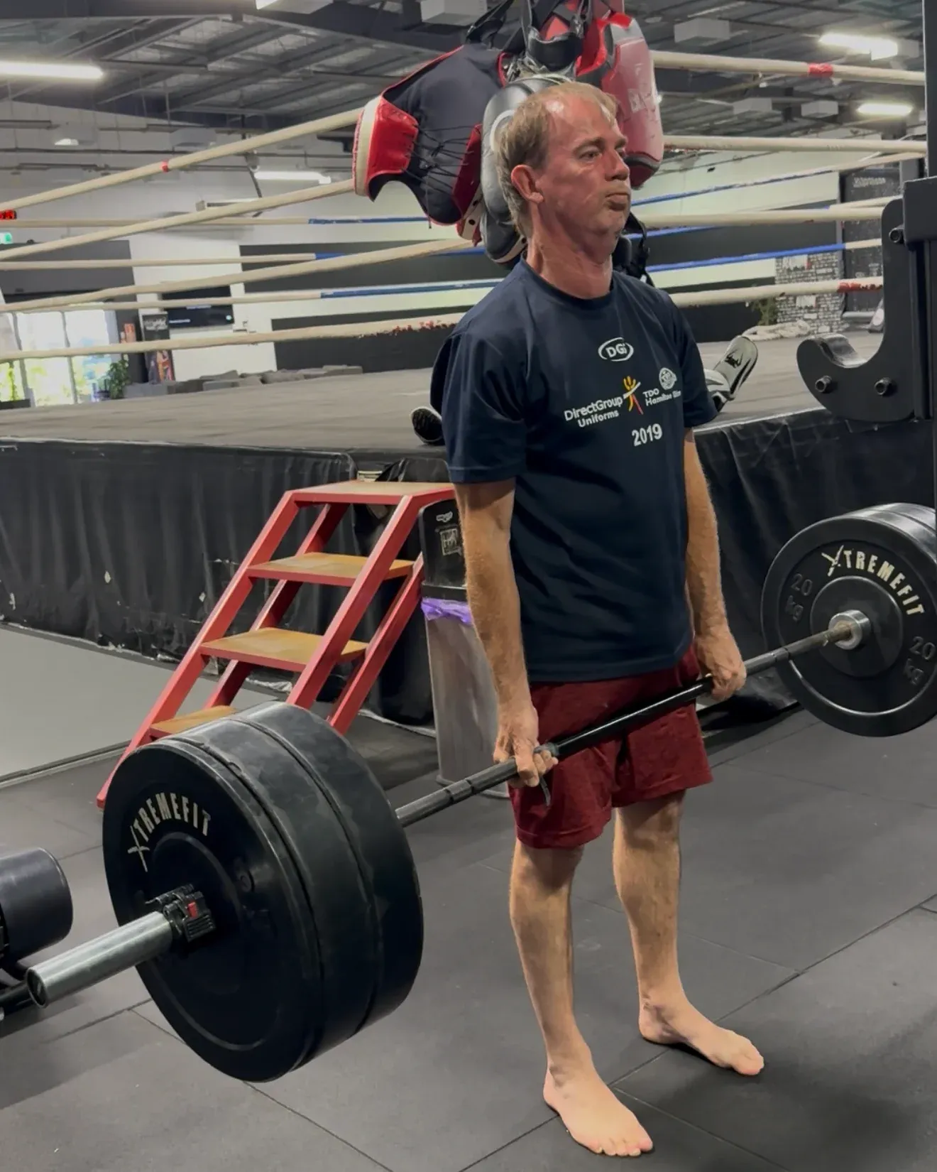 An older member lifting a loaded barbell, the boxing ring behind him