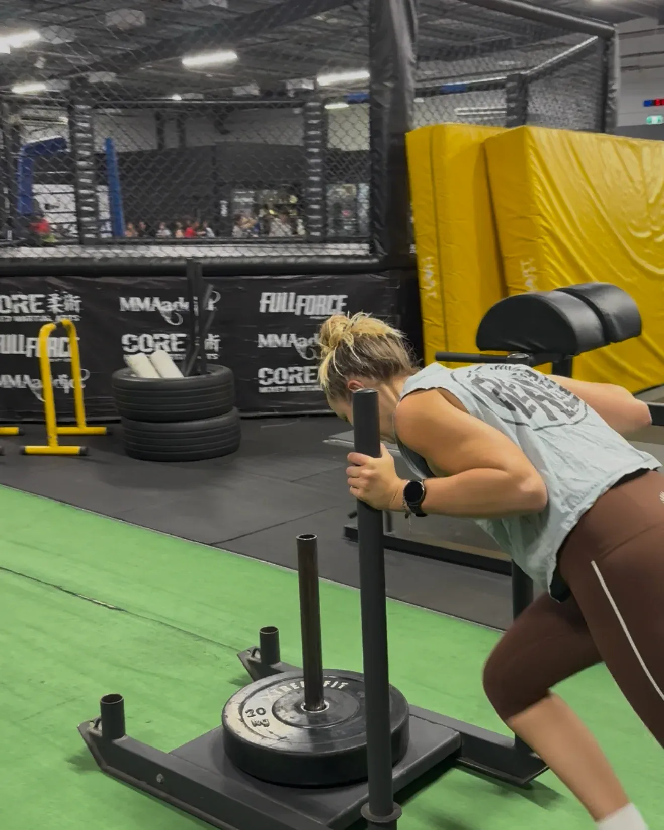 A member driving a weighted sled across the turf, the CORE MMA cage behind her